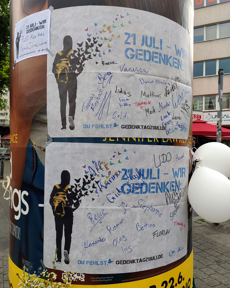 Mehrere weiße Plakate und Zettel sind an eine Lifaß-Säule geklebt. Anwesende des Gedenktags am Kottbusser-Tor haben darauf die Namen von vorzeitig Verstorbenen geschrieben, die sie vermissen.