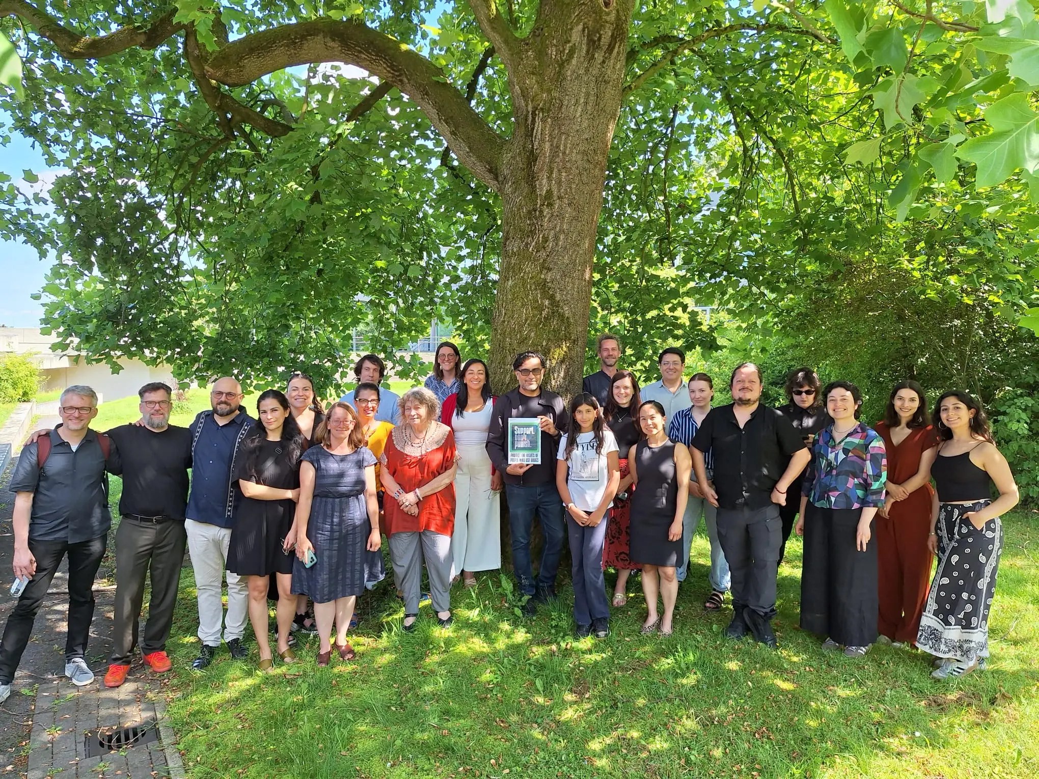 Gruppenfoto mit 23 Personen unter einem großen Baum. Die Person in der Mitte, Prof. Oswaldo Zavala, hält ein ausgedrucktes Schild mit dem Logo "Support Dont Punish"