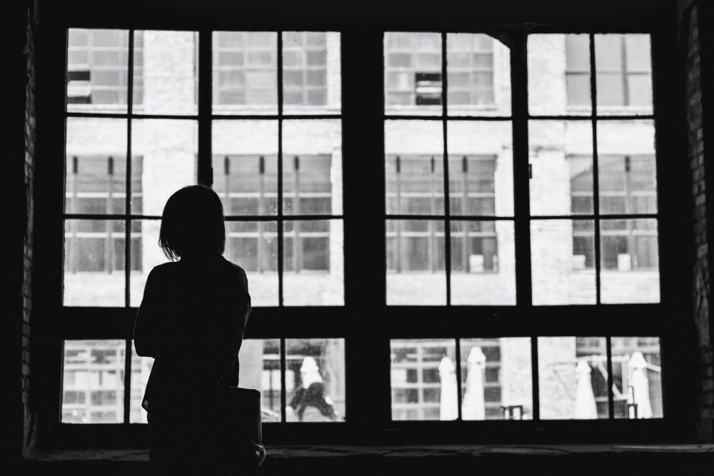 Eine Person steht am Fenster, mit dem Rücken zur Kamera. Wegen des Schattens sind nur Umrisse zu sehen. Es ist schwarz-weiß, wirkt anonym und einsam.
